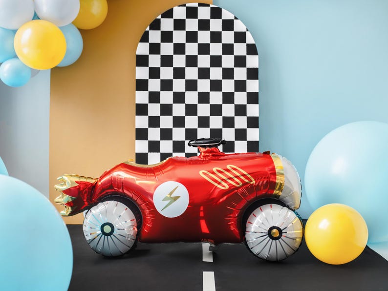 May include: A red race car shaped foil balloon with a lightning bolt design and gold accents. The balloon is in front of a black and white checkered background.