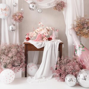 May include: A bridal shower party setup with a white backdrop, pink and white flowers, a table with a pink floral balloon, and silver disco balls.
