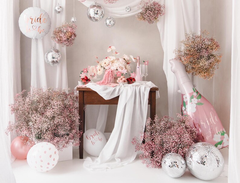 May include: A bridal shower party setup with a white backdrop, pink and white flowers, a table with a pink floral balloon, and silver disco balls.