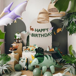 May include: A green dinosaur balloon, a purple pterodactyl balloon, and a green and brown dinosaur banner with the text "Happy Birthday" are displayed in a jungle-themed party setting. The scene includes a cake, cupcakes, and other decorations.