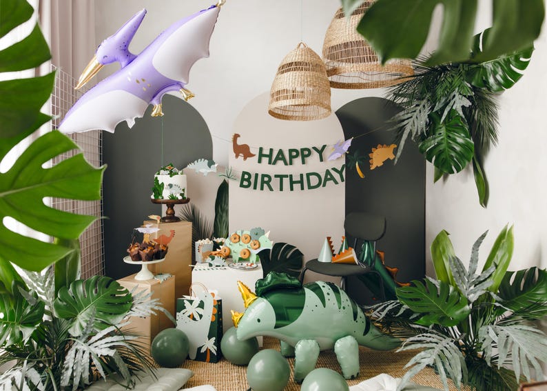 May include: A green dinosaur balloon, a purple pterodactyl balloon, and a green and brown dinosaur banner with the text "Happy Birthday" are displayed in a jungle-themed party setting. The scene includes a cake, cupcakes, and other decorations.