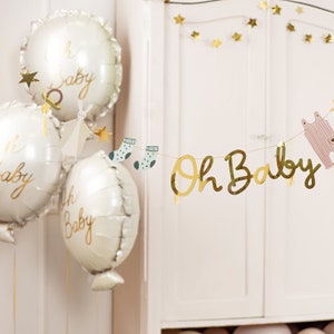 May include: A white baby shower banner with gold letters that read "Oh Baby" and gold stars. The banner is decorated with small paper cutouts of baby socks and a baby onesie. There are also three white balloons with gold lettering that read "Oh Baby" in the background.
