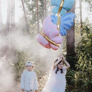May include: Two children, a boy and a girl, are holding a bunch of butterfly-shaped balloons in a forest setting. The balloons are pink, blue, and gold. The girl is wearing a white dress and the boy is wearing a white suit.