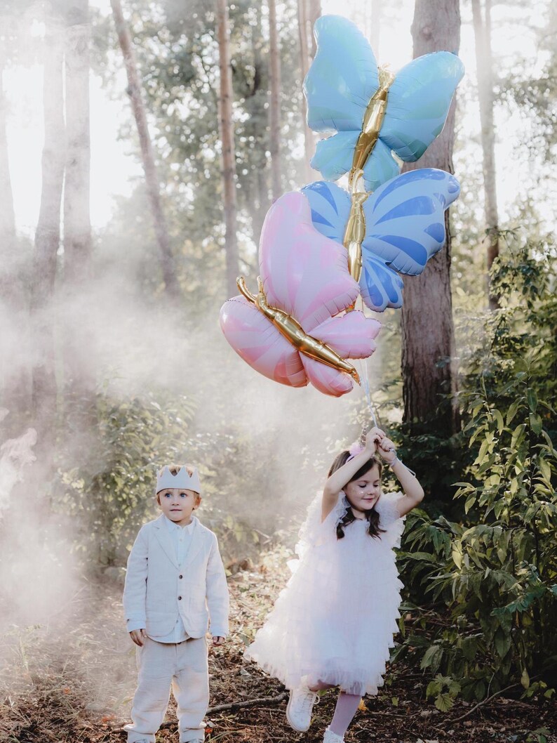 May include: Two children, a boy and a girl, are holding a bunch of butterfly-shaped balloons in a forest setting. The balloons are pink, blue, and gold. The girl is wearing a white dress and the boy is wearing a white suit.