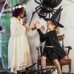 Op de afbeelding: Twee kinderen verkleed in Halloween-kostuums. E&eacute;n kind draagt een witte jurk en een bloemenkrans. Het andere kind draagt een zwart heksenkostuum met een zwarte hoed en glimmende panty's. Ze staan voor een grote zwarte spinballon.