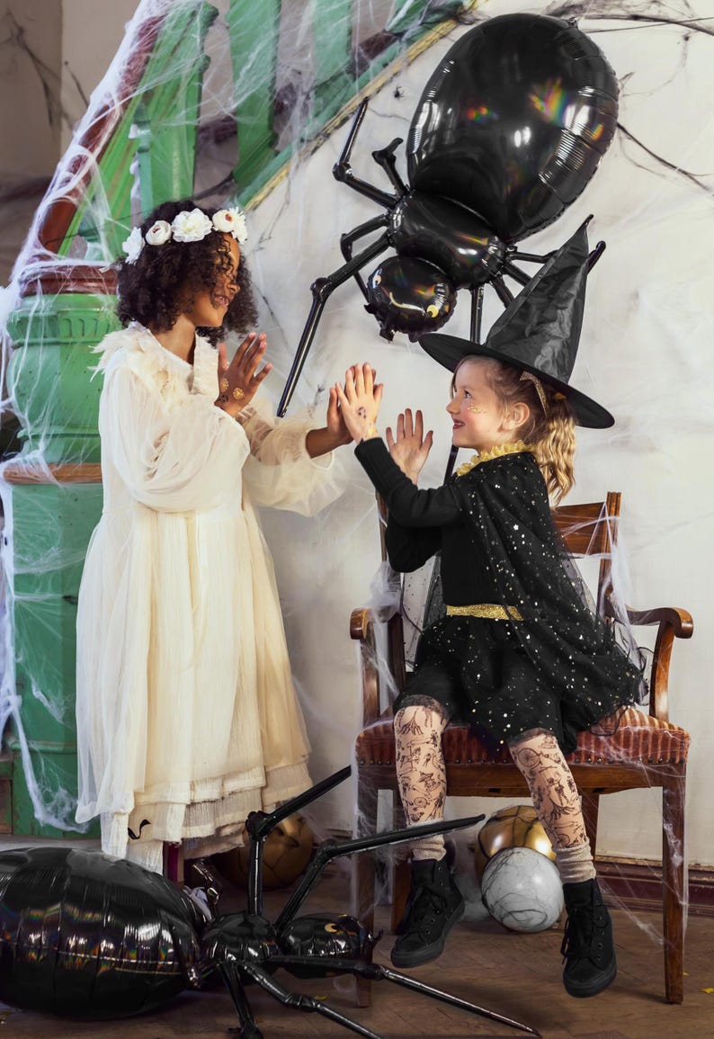 Op de afbeelding: Twee kinderen verkleed in Halloween-kostuums. E&eacute;n kind draagt een witte jurk en een bloemenkrans. Het andere kind draagt een zwart heksenkostuum met een zwarte hoed en glimmende panty's. Ze staan voor een grote zwarte spinballon.