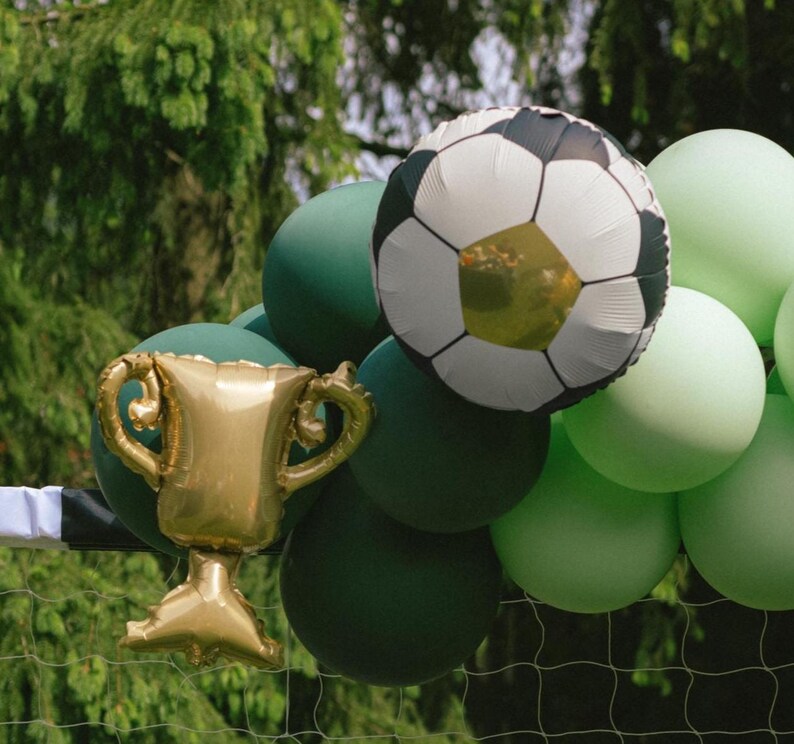 May include: A gold trophy balloon and a soccer ball balloon are surrounded by green and light green balloons. The balloons are arranged in front of a green background, possibly a soccer net.