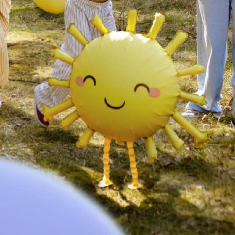 May include: A yellow sun-shaped balloon with a smiling face and rosy cheeks. The balloon has orange and yellow striped legs.