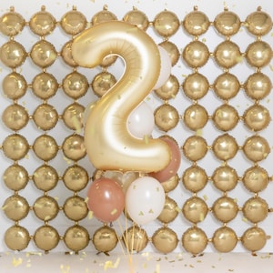 May include: A gold number two balloon with a gold balloon backdrop. The number two balloon is surrounded by smaller balloons in gold, white, and brown.