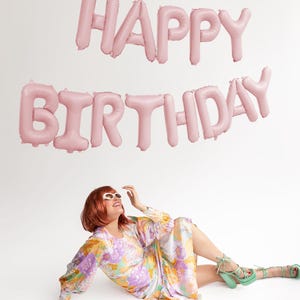 May include: Pink foil balloons spelling out "Happy Birthday" against a white background. A person wearing a floral dress and sunglasses is sitting on the floor looking up at the balloons.