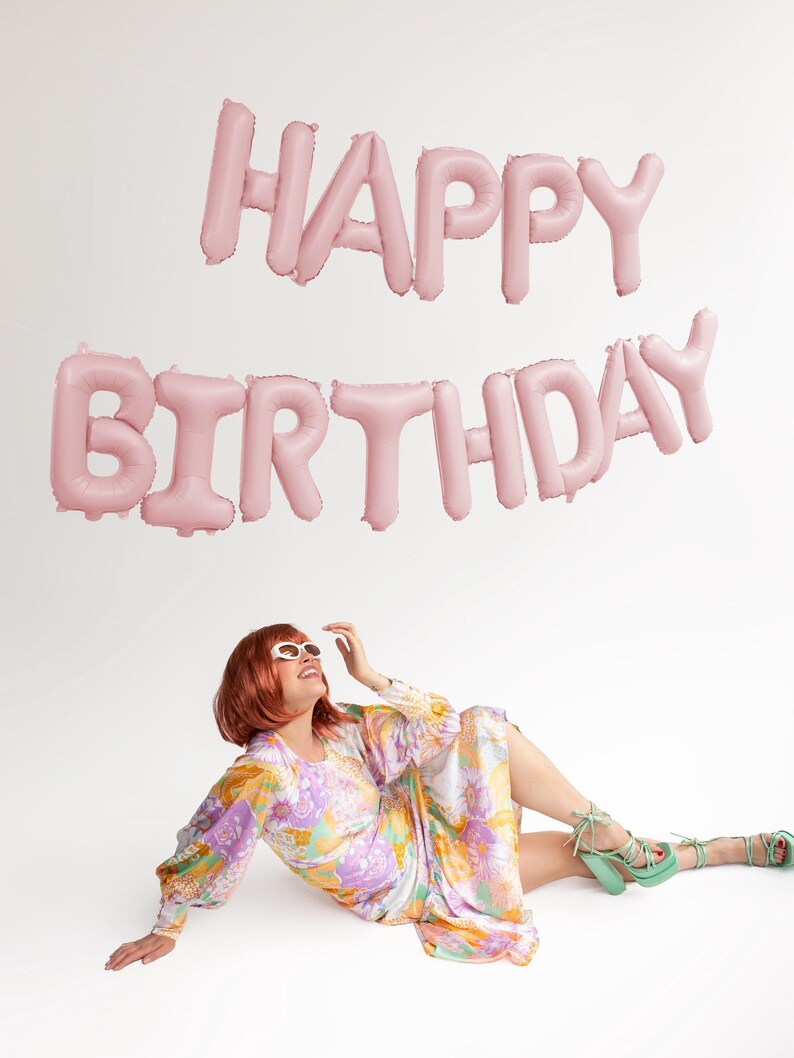May include: Pink foil balloons spelling out "Happy Birthday" against a white background. A person wearing a floral dress and sunglasses is sitting on the floor looking up at the balloons.