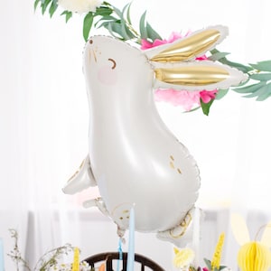 A cute white bunny with golden ears. The balloon is surrounded by colorful flowers, paper decorations, candles, and greenery, creating a cheerful and whimsical spring or Easter celebration atmosphere.
