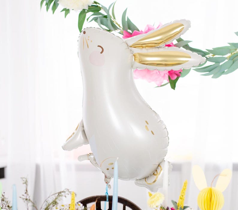 A cute white bunny with golden ears. The balloon is surrounded by colorful flowers, paper decorations, candles, and greenery, creating a cheerful and whimsical spring or Easter celebration atmosphere.