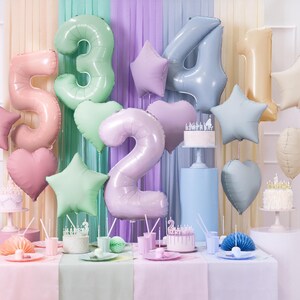 May include: A festive birthday party scene with pastel-colored balloons shaped like numbers, stars, and hearts. The balloons are in shades of pink, green, and blue. Small cakes with candles and party supplies are on the table.