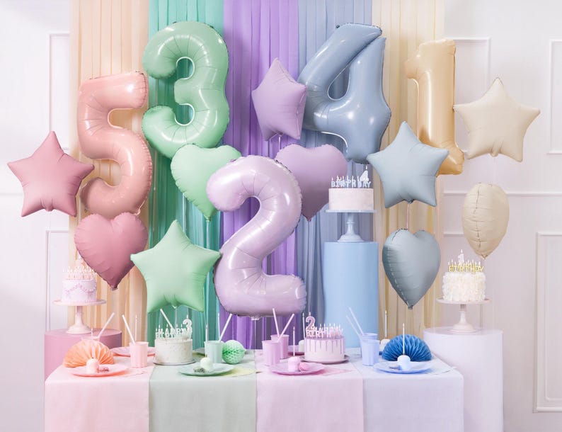 May include: A festive birthday party scene with pastel-colored balloons shaped like numbers, stars, and hearts. The balloons are in shades of pink, green, and blue. Small cakes with candles and party supplies are on the table.