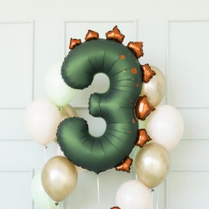 Könnte beinhalten: Ein grüner Folienballon mit der Zahl 3 und kupferfarbenen Zacken, umgeben von weißen, goldenen und hellgrünen Luftballons. Der Ballon mit der Zahl 3 hat eine strukturierte Oberfläche und ist als Dinosaurier gestaltet.