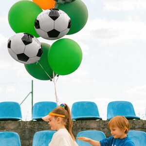 May include: A collection of colorful balloons, including green, orange, and soccer ball-themed designs, float against a cloudy sky. The balloons are held by a string. Two children are in the foreground, one holding a soccer ball.