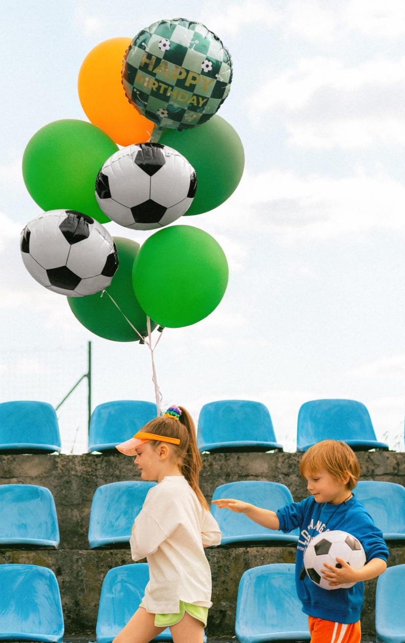 May include: A collection of colorful balloons, including green, orange, and soccer ball-themed designs, float against a cloudy sky. The balloons are held by a string. Two children are in the foreground, one holding a soccer ball.