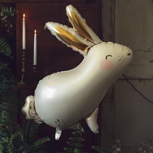 May include: A white balloon shaped like a rabbit with gold ears and a pink nose. The balloon is partially deflated and has a textured surface.