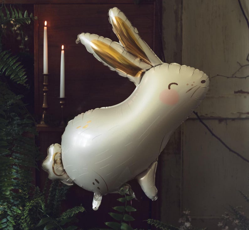 May include: A white balloon shaped like a rabbit with gold ears and a pink nose. The balloon is partially deflated and has a textured surface.