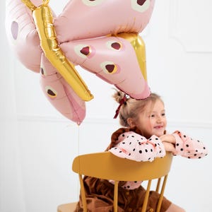 May include: A young girl sits on a yellow chair holding a pink and gold butterfly balloon. The balloon has a red and white polka dot pattern.