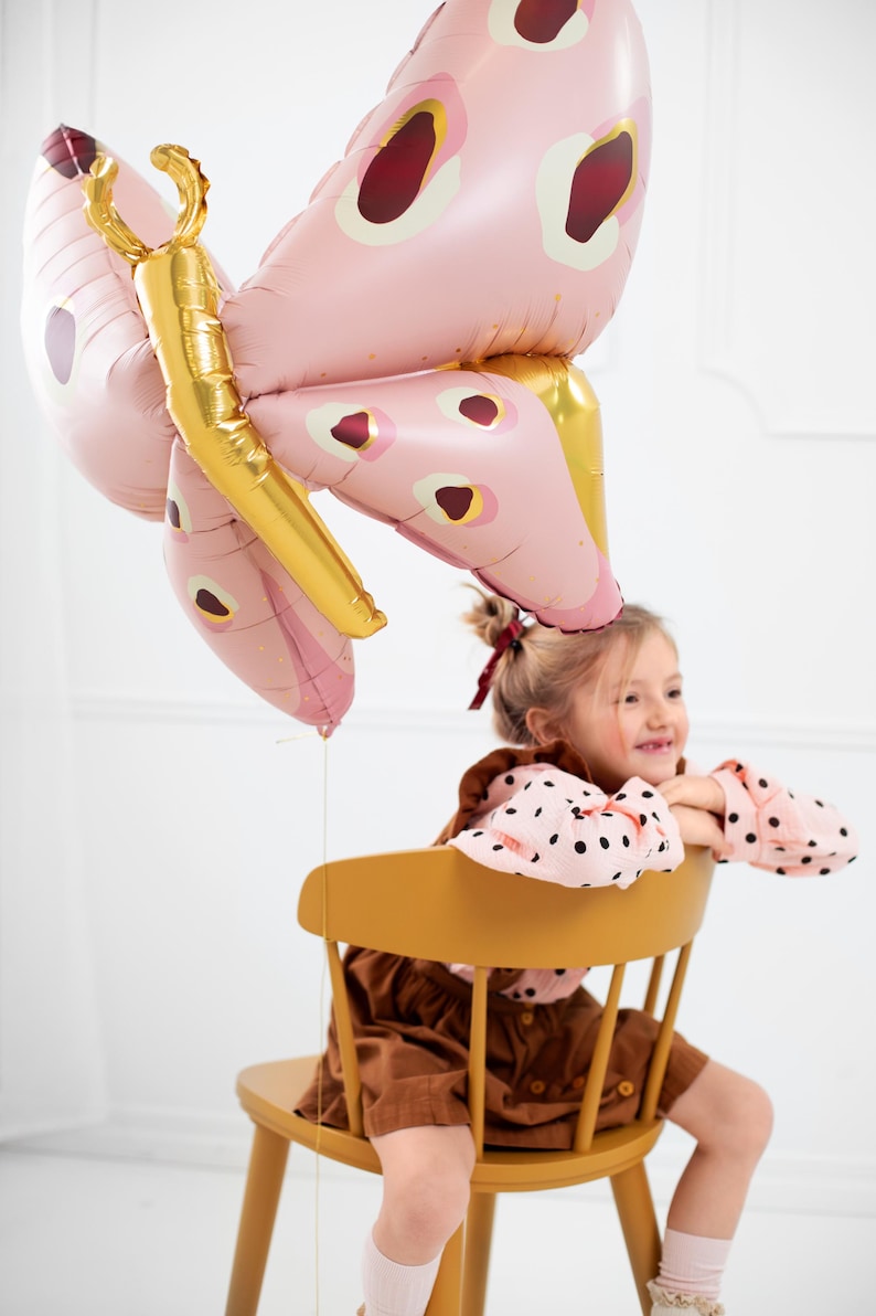 May include: A young girl sits on a yellow chair holding a pink and gold butterfly balloon. The balloon has a red and white polka dot pattern.