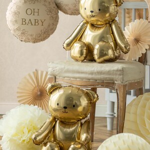 May include: Two gold teddy bear-shaped balloons are featured in this image, along with two round balloons that read "OH BABY". The balloons are surrounded by paper decorations in shades of cream and gold, creating a festive atmosphere.