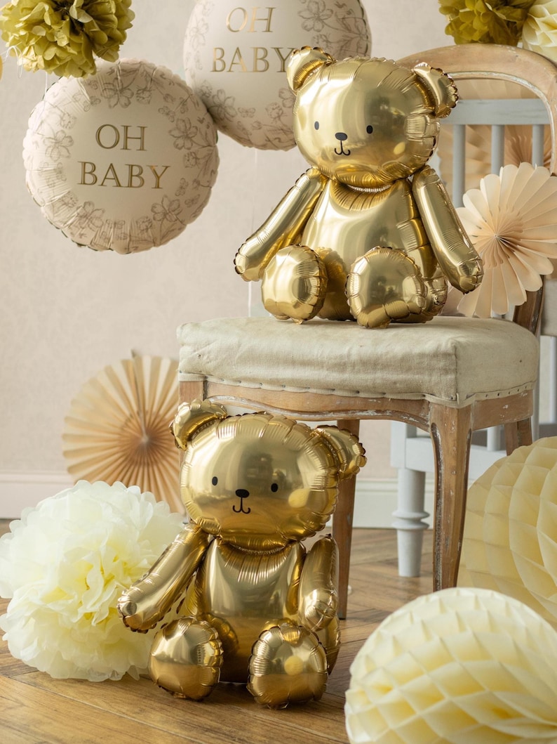May include: Two gold teddy bear-shaped balloons are featured in this image, along with two round balloons that read "OH BABY". The balloons are surrounded by paper decorations in shades of cream and gold, creating a festive atmosphere.