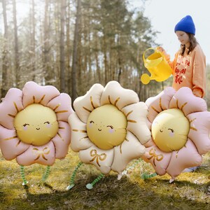 Whimsical outdoor scene with a child holding a yellow watering can beside three adorable flower-shaped foil balloons with smiling faces, pink and cream petals, perfect for spring parties, baby showers, and garden-themed birthday celebrations.