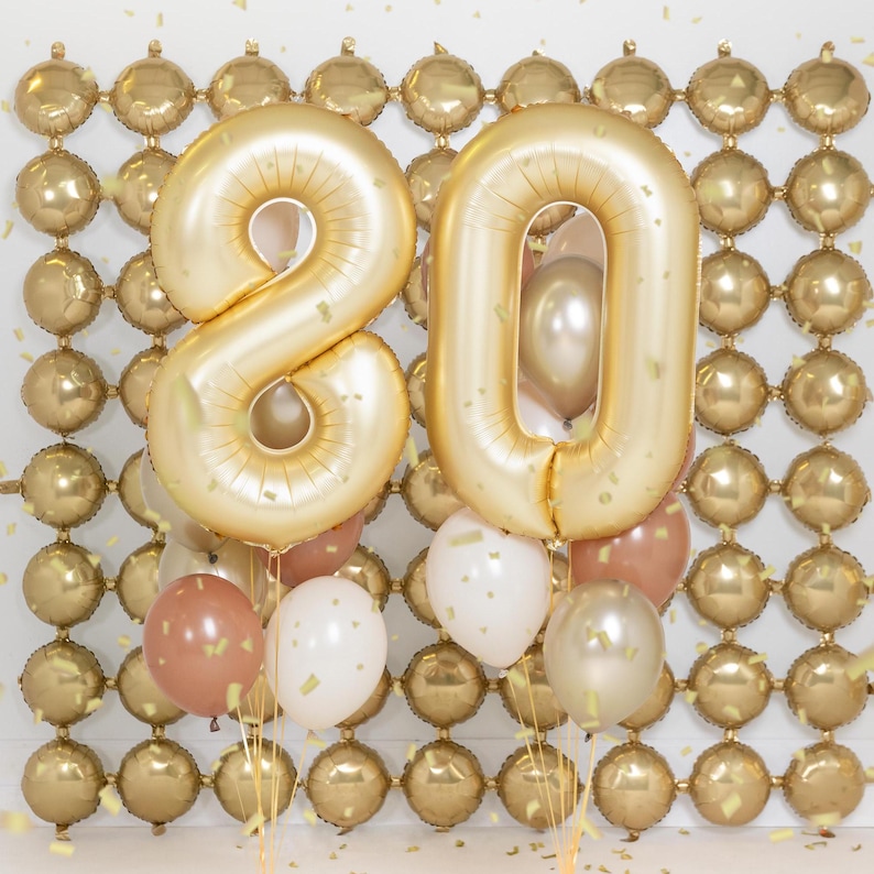 May include: Gold number balloons spelling out "80" with smaller gold and brown balloons surrounding them. The balloons are in front of a wall of smaller gold balloons.