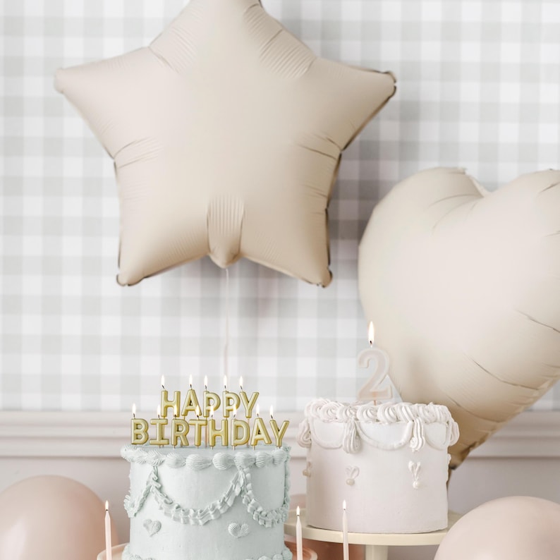Puede incluir: Dos pasteles decorados con glaseado y velas. Un pastel tiene una vela con la inscripci&oacute;n "Feliz cumplea&ntilde;os" y el otro tiene una vela con el n&uacute;mero "2". Los pasteles est&aacute;n sobre una mesa con un mantel a cuadros blanco y gris. Hay dos globos en el fondo, uno con forma de estrella y el otro con forma de coraz&oacute;n.