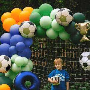 May include: A colorful balloon arch in shades of orange, blue, green, and white, decorated with soccer ball and trophy balloons. A young person in a blue shirt holds a popcorn container in front of a soccer net.