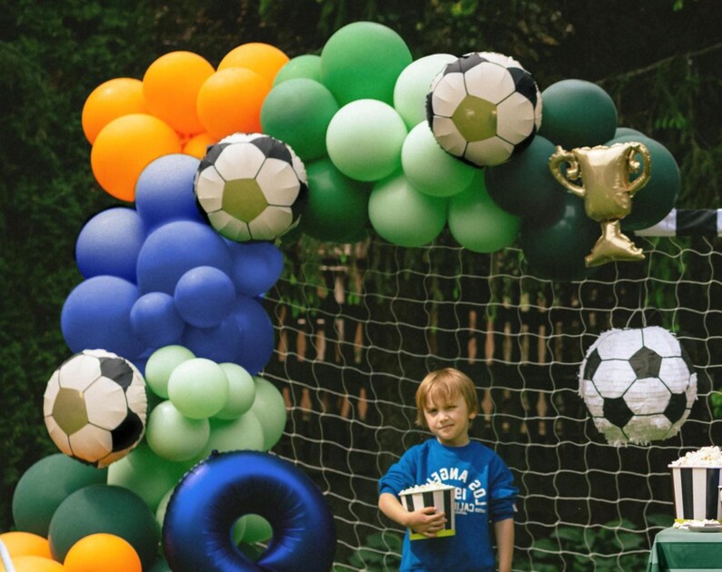 May include: A colorful balloon arch in shades of orange, blue, green, and white, decorated with soccer ball and trophy balloons. A young person in a blue shirt holds a popcorn container in front of a soccer net.
