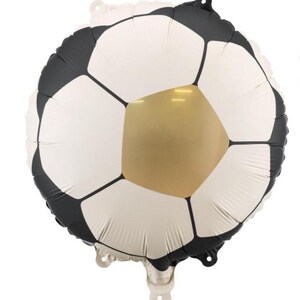 May include: A soccer ball-shaped foil balloon. The balloon features a classic soccer ball design with white pentagons, black hexagons, and a gold-colored pentagon in the center. It has a shiny, metallic finish and is designed for party decorations.
