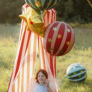 May include: A young person sits in a striped tent, surrounded by festive balloons. The balloons include a gold star-shaped balloon, a red and white striped balloon with gold stars, and a blue and green striped balloon with gold stars. The tent has red and white stripes.