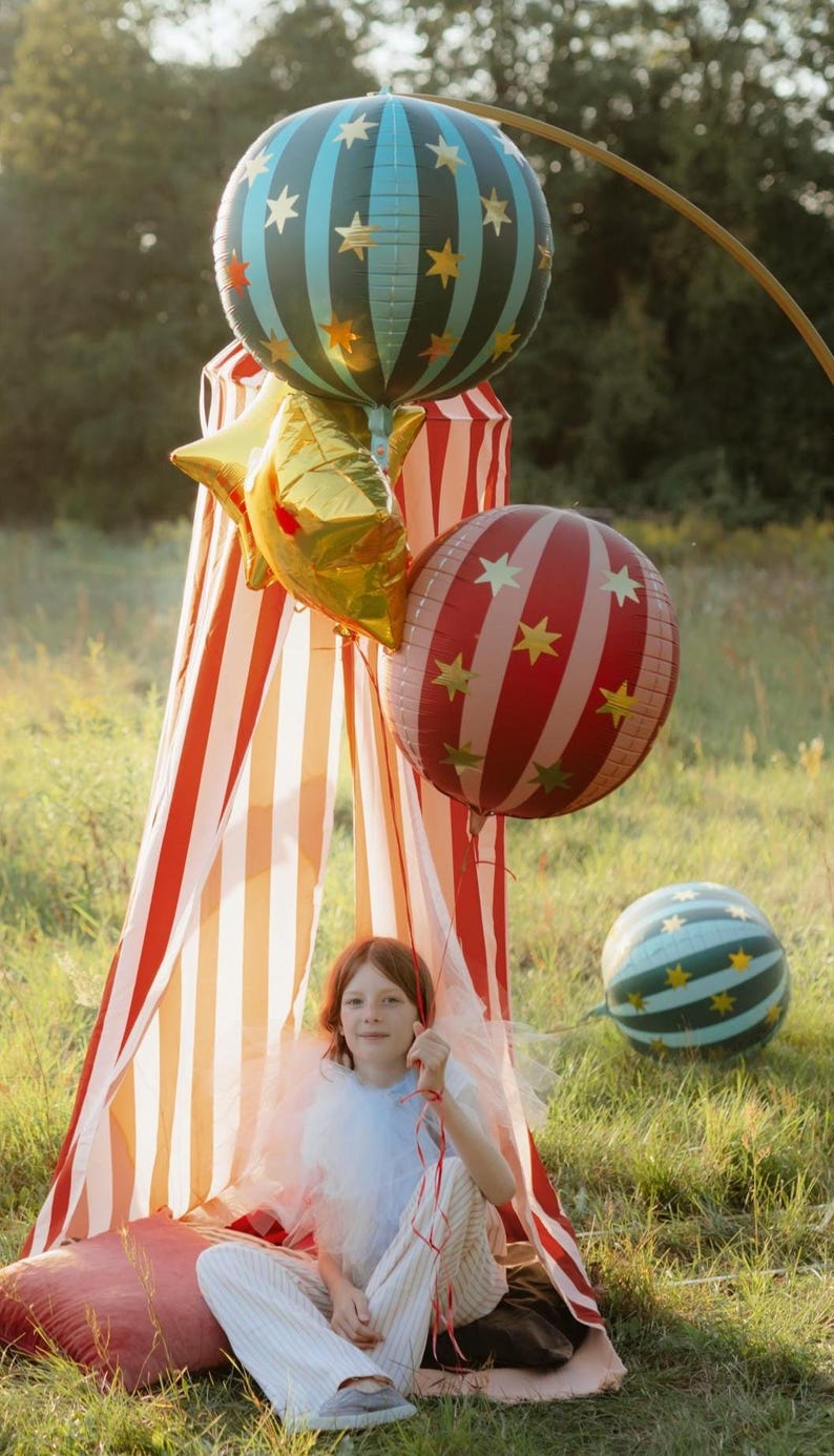 May include: A young person sits in a striped tent, surrounded by festive balloons. The balloons include a gold star-shaped balloon, a red and white striped balloon with gold stars, and a blue and green striped balloon with gold stars. The tent has red and white stripes.