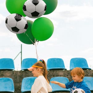 May include: A collection of balloons, including soccer ball and green balloons, and a Happy Birthday balloon. Two children are walking on stadium steps. One child is holding a soccer ball. The sky is visible in the background.