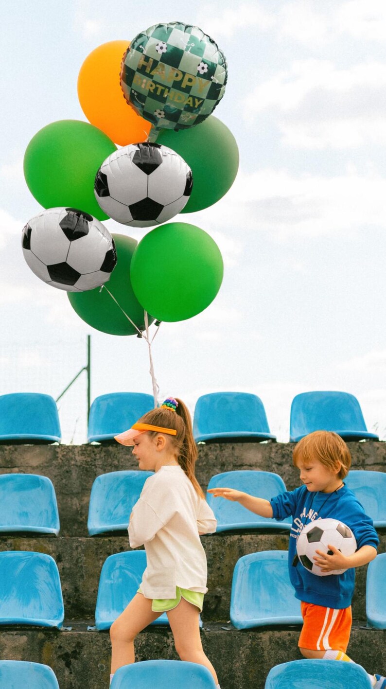 May include: A collection of balloons, including soccer ball and green balloons, and a Happy Birthday balloon. Two children are walking on stadium steps. One child is holding a soccer ball. The sky is visible in the background.