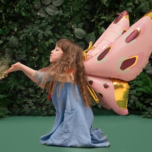 May include: A young girl wearing a blue dress and a pink and gold butterfly costume blows glitter from her hand. The background is a green wall with lush foliage.