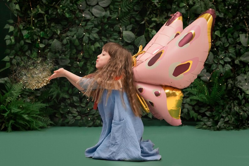 May include: A young girl wearing a blue dress and a pink and gold butterfly costume blows glitter from her hand. The background is a green wall with lush foliage.