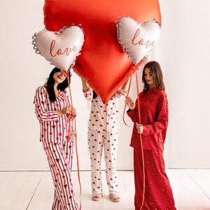 May include: Three people wearing red and white pajamas hold large red heart-shaped balloons with the word "love" written on them.