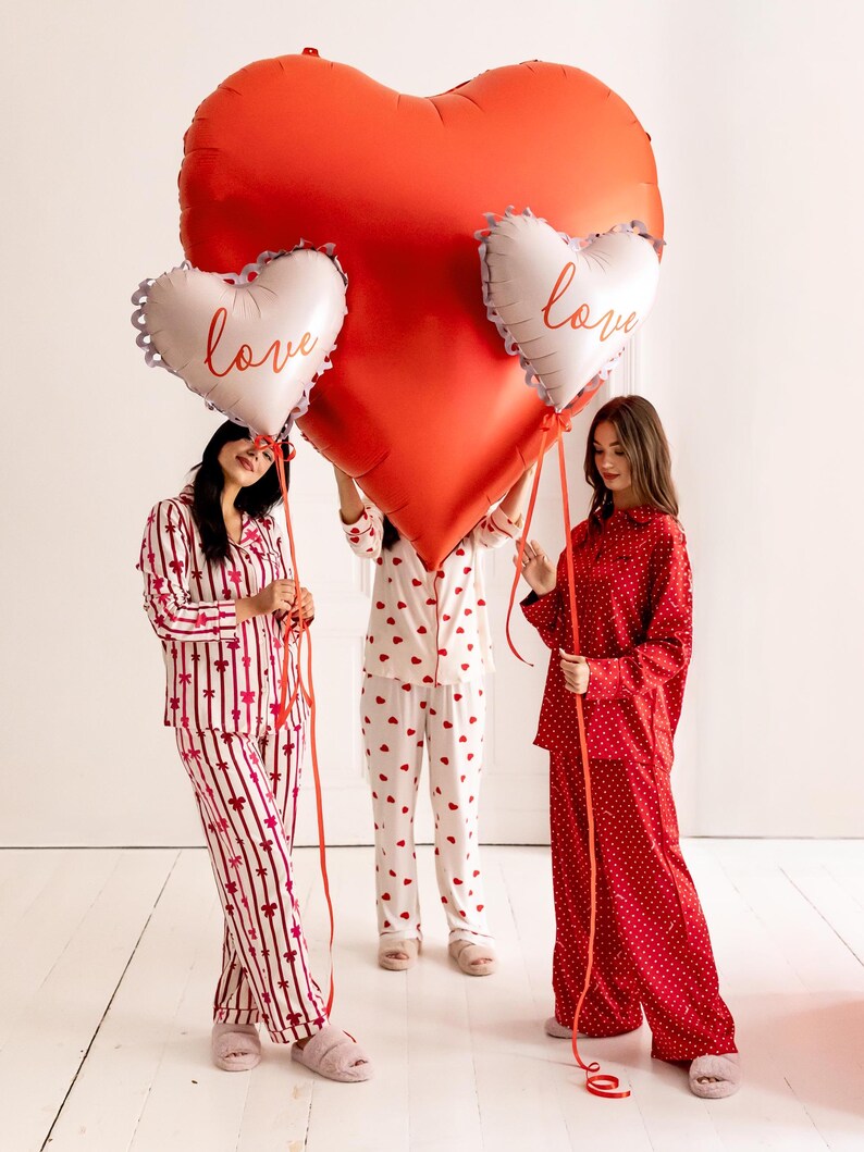 May include: Three people wearing red and white pajamas hold large red heart-shaped balloons with the word "love" written on them.