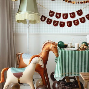 Puede incluir: Una escena de fiesta de cumplea&ntilde;os con globos con forma de caballo, una pancarta de "Feliz Cumplea&ntilde;os" y botas de vaquero. Los globos son en tonos marrones y beige, y la mesa est&aacute; cubierta con un mantel a rayas verdes y blancas.