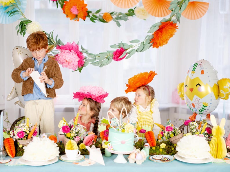 Op de afbeelding: Een feestelijke Paassc&egrave;ne met kinderen en decoraties. Een jongen in een bruine trui en kinderen met bloemenhoeden verzamelen zich rond een tafel met een taart, bloemen en een kuikenvormige ballon. De tafel is versierd met wortels en papieren decoraties.