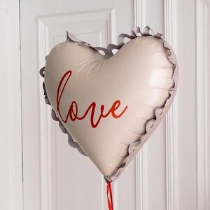 Heart-shaped foil balloon with the word "love" written in red script, featuring a ruffled edge. Ideal for romantic occasions like Valentine's Day, anniversaries, or weddings.