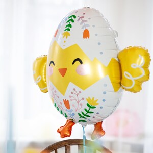 Op de afbeelding: Een vrolijke paasballon in de vorm van een uitkomend kuiken. De ballon heeft een geel en wit eierontwerp met bloemaccenten, een lachend kuikengezicht en gele vleugelvormige bevestigingen. Oranje voeten bungelen eronder.