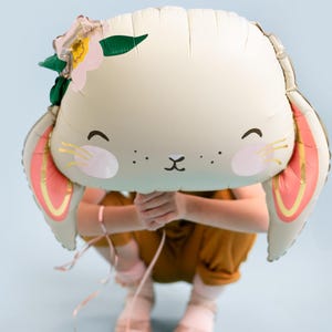 May include: A white bunny balloon with pink ears and a flower crown. The balloon is held in front of a person wearing a brown jumpsuit.