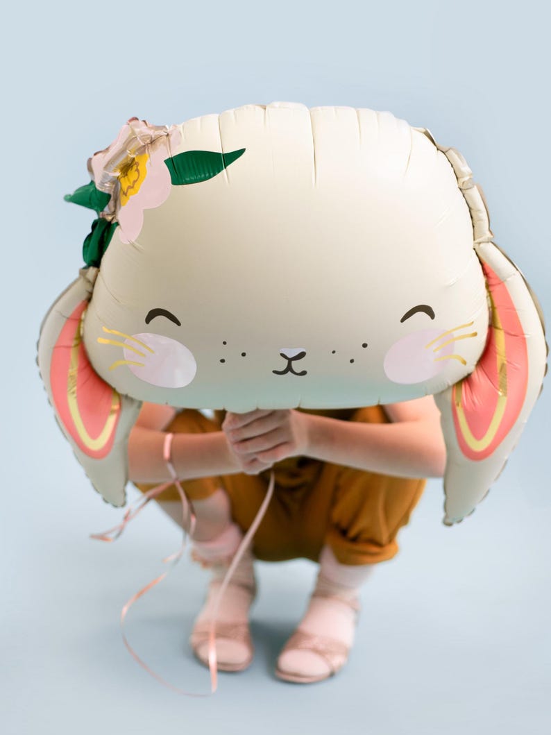 May include: A white bunny balloon with pink ears and a flower crown. The balloon is held in front of a person wearing a brown jumpsuit.