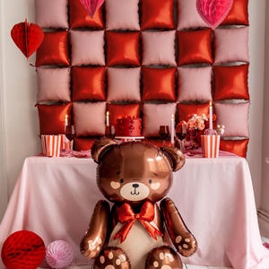 Pu&ograve; includere: Un palloncino a forma di orsacchiotto marrone con un fiocco rosso si trova su una tovaglia rosa davanti a un muro decorato con decorazioni a forma di cuore rosse e rosa.