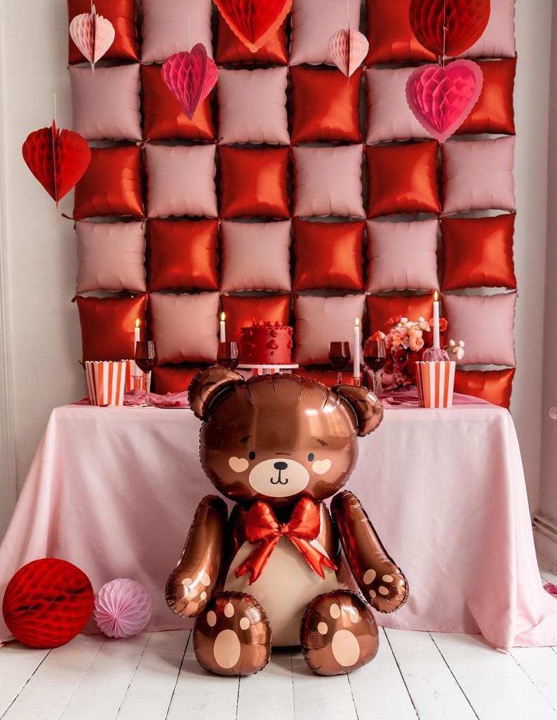 Pu&ograve; includere: Un palloncino a forma di orsacchiotto marrone con un fiocco rosso si trova su una tovaglia rosa davanti a un muro decorato con decorazioni a forma di cuore rosse e rosa.